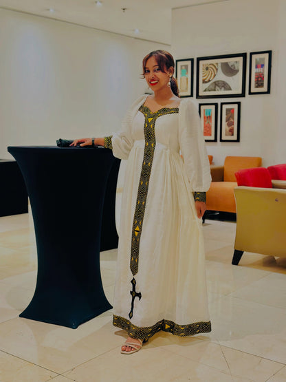 A woman wearing a long white traditional Ethiopian shop near me with green and gold embroidery, standing next to a black podium in a modern indoor lobby setting.