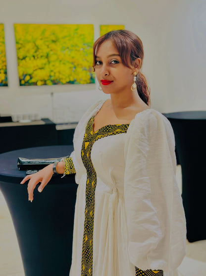 A woman wearing a white traditional Ethiopian shop near me with green and gold patterned trim, standing indoors near a black table.