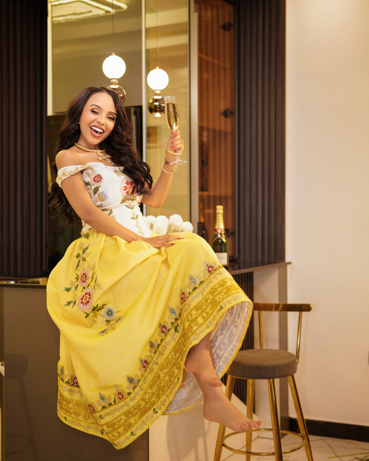 A woman sitting on a bar stool in a restaurant or lounge setting, wearing a bright yellow off-the-shoulder Habesha Kemis with floral embroidery and holding a glass of champagne.