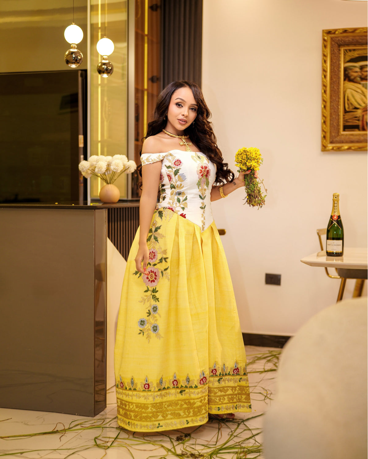 A woman wearing a bright yellow off-the-shoulder Habesha Kemis with a white embroidered bodice and floral details, holding a bouquet of yellow flowers in an indoor setting.