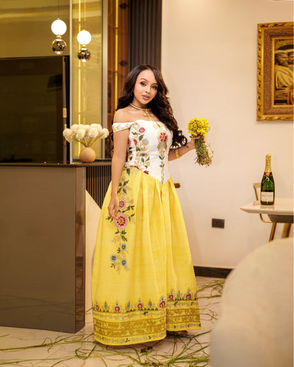 A woman wearing a bright yellow off-the-shoulder Habesha Kemis with a white embroidered bodice and floral details, holding a bouquet of yellow flowers in an indoor setting.