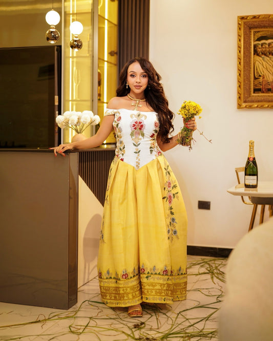 A woman models a yellow off-the-shoulder maxi Habesha Kemis with a white embroidered bodice and floral details, holding a bouquet of yellow flowers in an indoor setting.