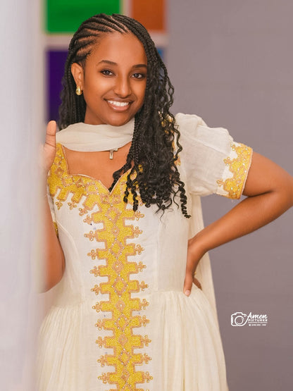 A smiling woman with long black braided hair wearing a cream-colored traditional Ethiopian Dress with prominent gold embroidery down the front and on the sleeve cuffs, posed against a soft gray background with colorful blocks.