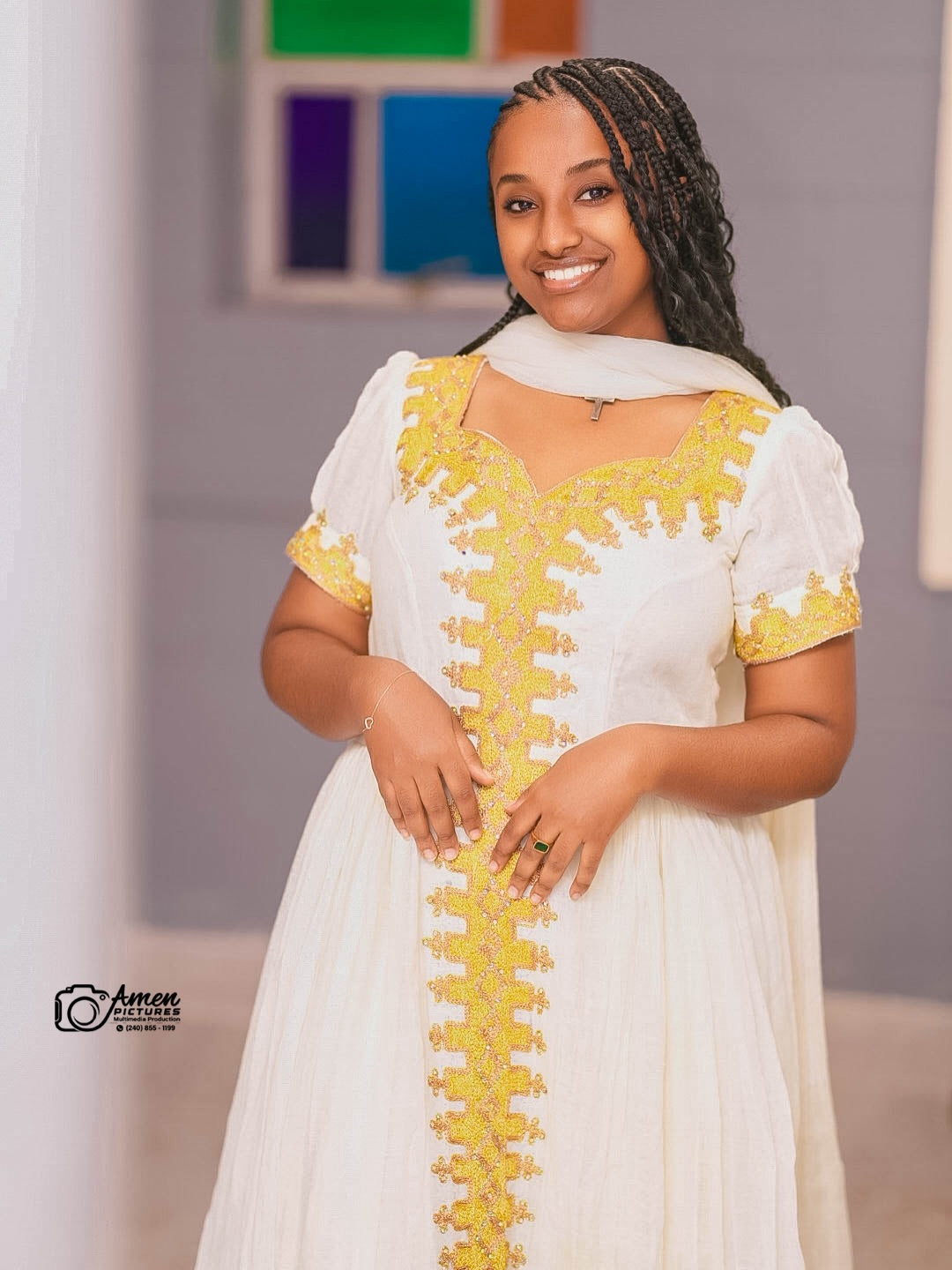 A smiling woman wearing a white traditional Ethiopian Dress with prominent yellow embroidery down the front and on the sleeves, paired with a white headscarf, set against a softly blurred indoor background.
