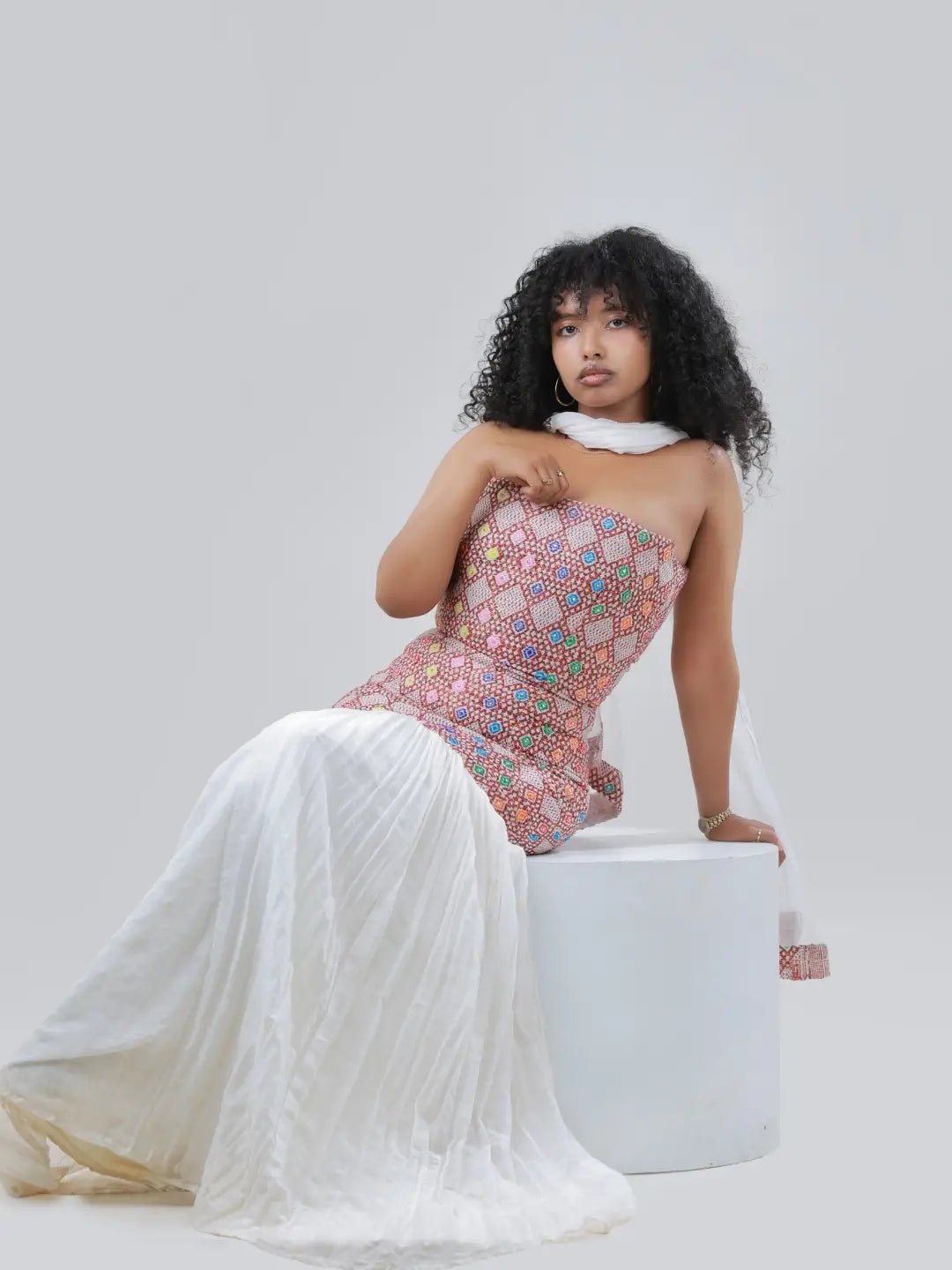 A model with curly black hair sits on a white Habesha Kemis, wearing a strapless fitted dress with a pink, green, and blue geometric pattern on the bodice and a flowing white skirt.