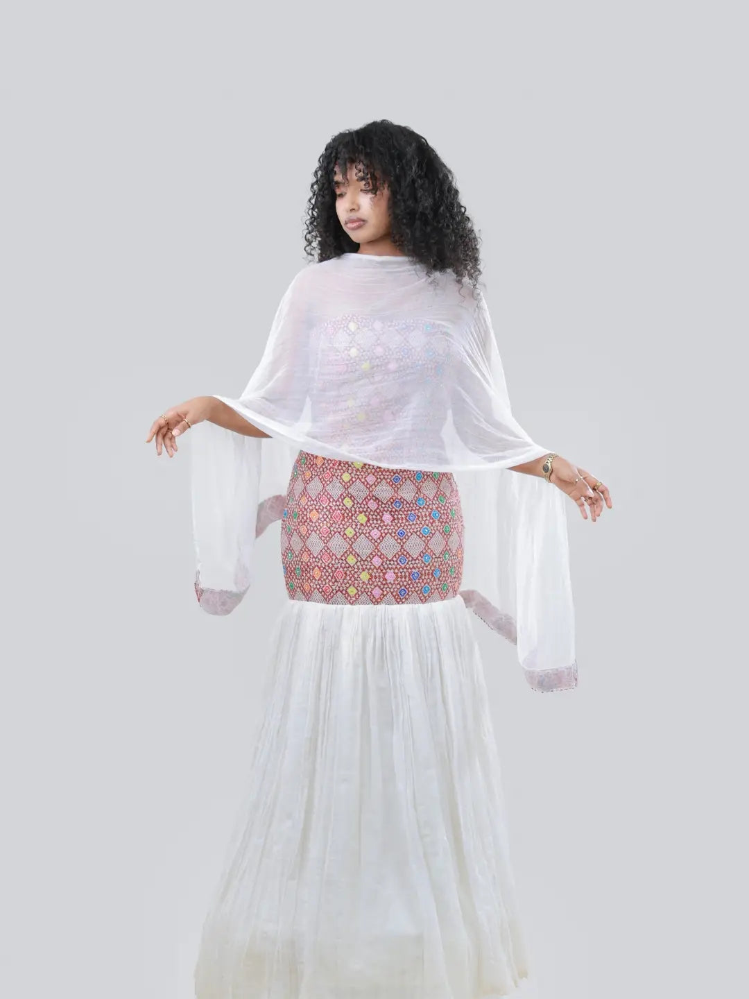 A woman modeling a white sheer long-sleeve tunic with wide bell sleeves, worn over a pink and multicolor patterned embroidered Habesha Kemis, against a solid gray studio background.