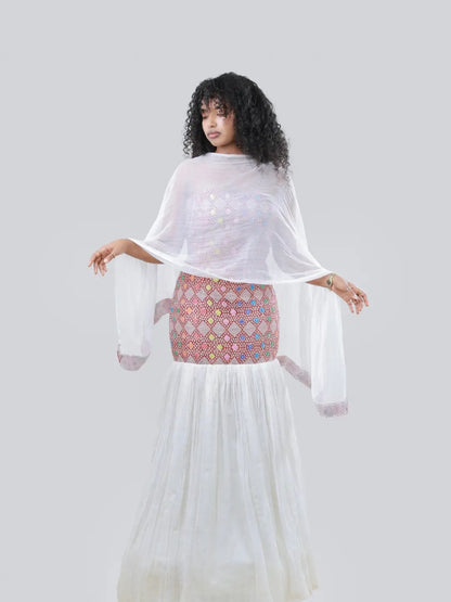 A woman modeling a white sheer long-sleeve tunic with wide bell sleeves, worn over a pink and multicolor patterned embroidered Habesha Kemis, against a solid gray studio background.