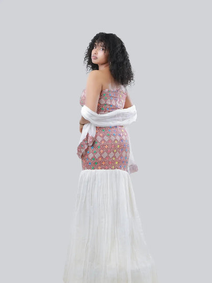 A woman wearing a strapless Habesha Kemis with a colorful geometric pattern on the bodice and a flowing white tulle skirt, posed against a solid light gray background.