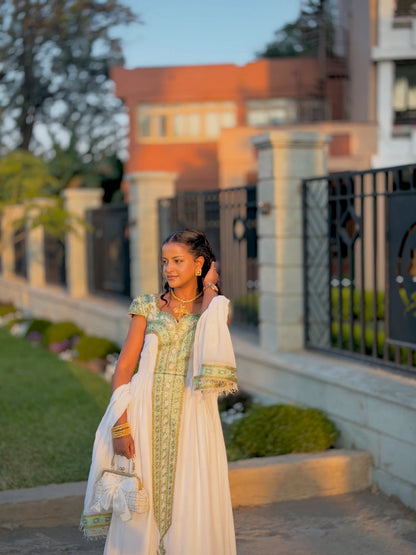 A woman wearing a traditional white and light green embroidered Habesha Store stands outdoors in front of a stone wall and black metal fence, holding a matching white shawl and a small white handbag.