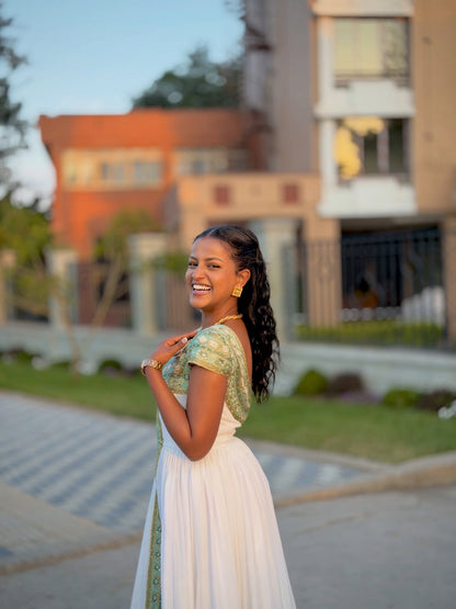 A woman wearing a white and green traditional Habesha Store stands outdoors on a paved walkway with blurred buildings and greenery in the background.