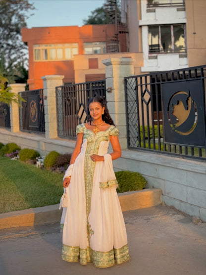 A woman wearing a traditional Habesha Store in off-white with green and gold embroidery, standing outdoors in front of a modern building and black metal fence.