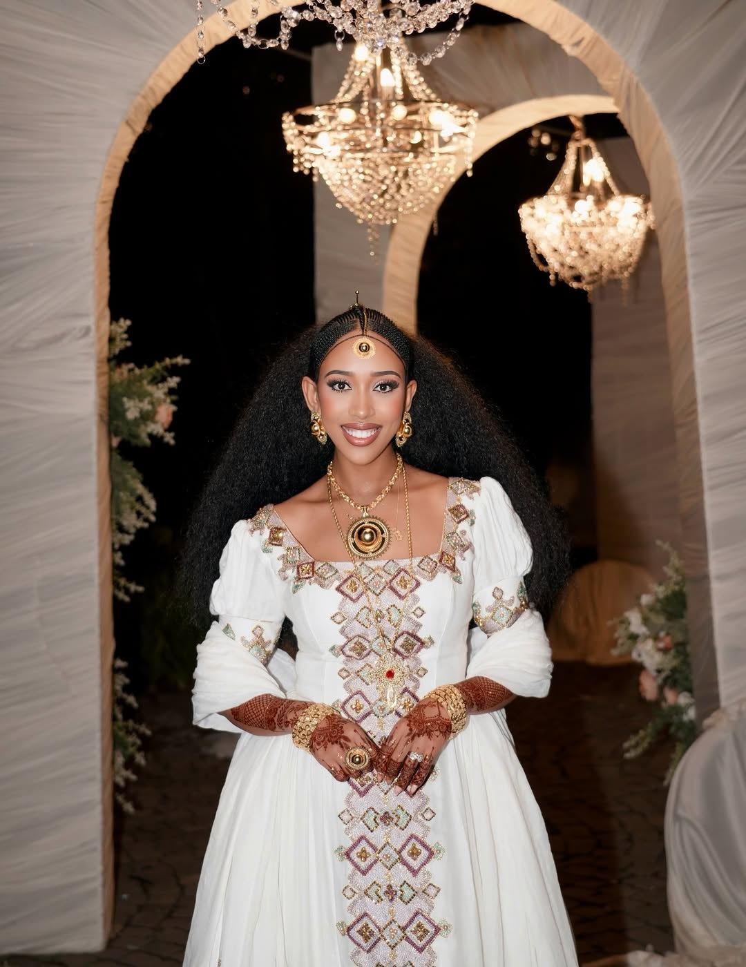 Woman in a white embroidered dress standing in front of a decorative arch with chandeliers.