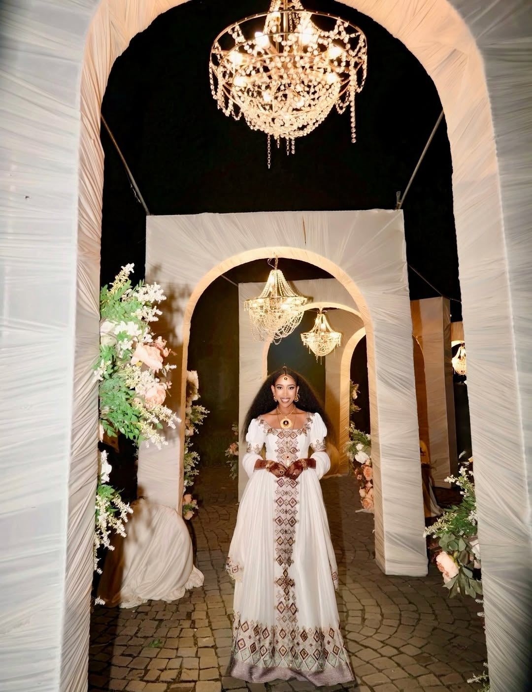 Woman in a white dress standing in an archway with chandeliers and floral decorations.