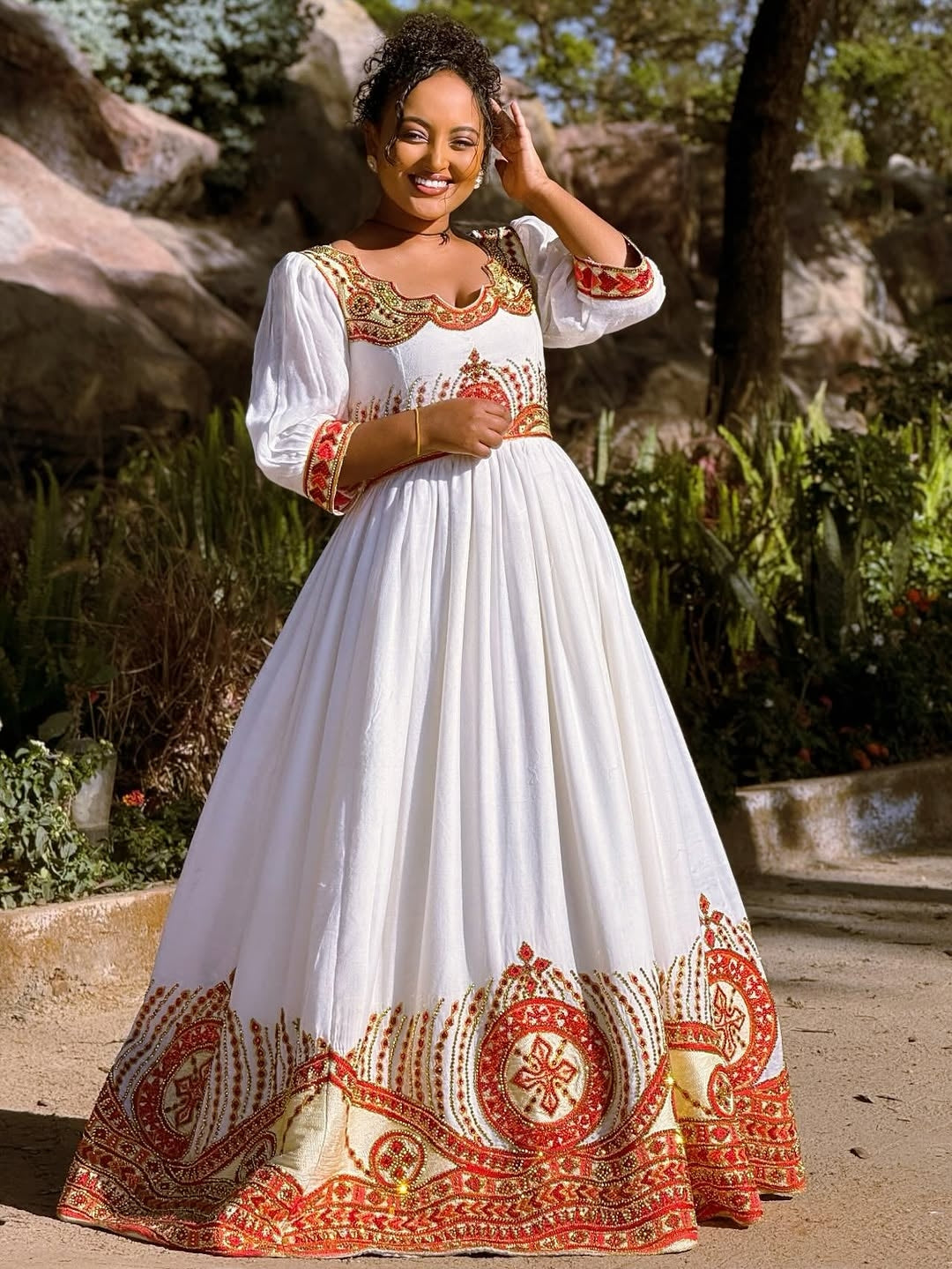 Woman in a white Ethiopian cultural dress   with red and gold embroidery standing outdoors.