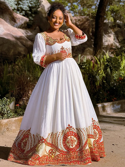 Woman in a white Ethiopian cultural dress   with red and gold embroidery standing outdoors.