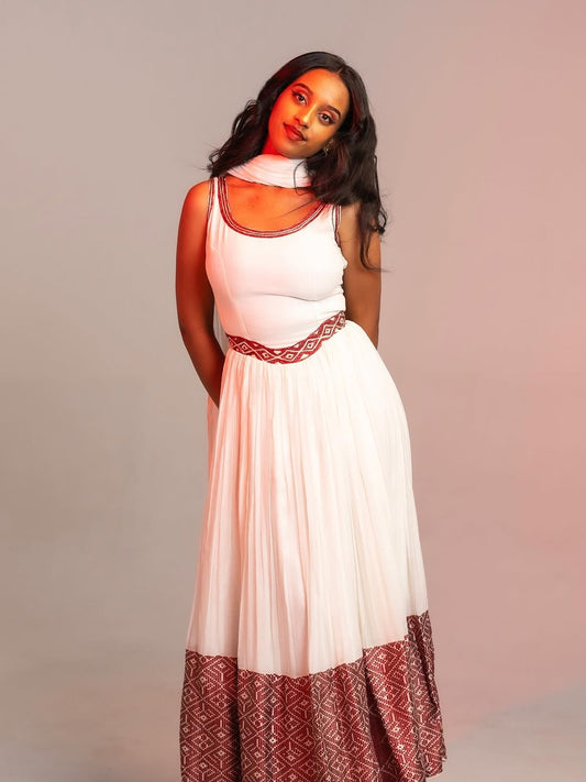 Woman wearing a white Ethiopian cultural dress with red patterned hem against a plain background