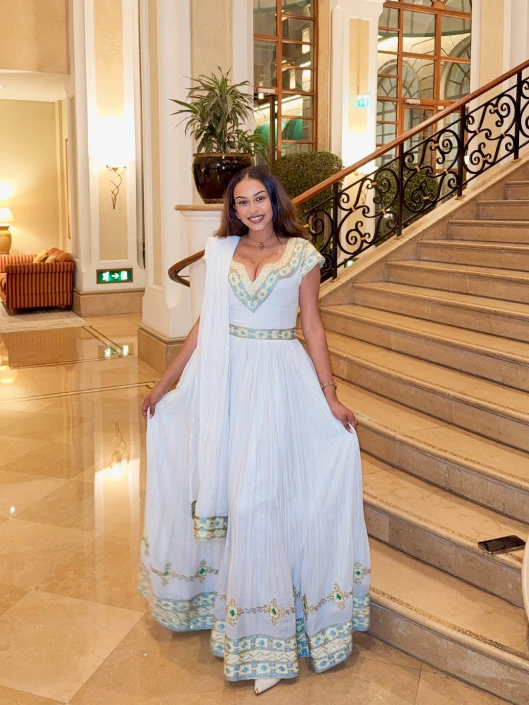Woman in a white Ethiopian dress with green accents standing on a staircase in an elegant interior setting.