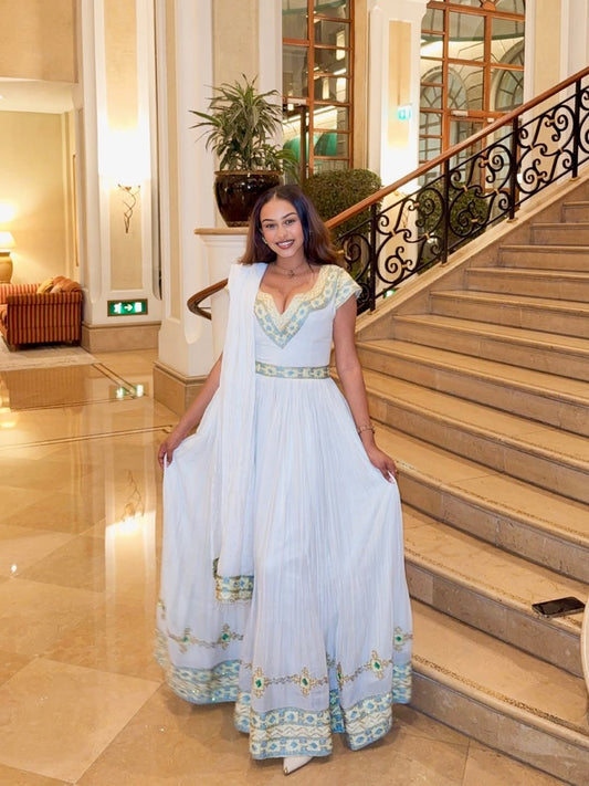 Woman in a white Ethiopian dress with green accents standing on a staircase in an elegant interior setting.