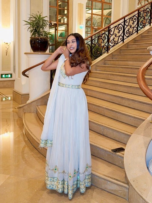 Woman in a white Ethiopian dress with green accents standing on a staircase in an elegant interior setting.