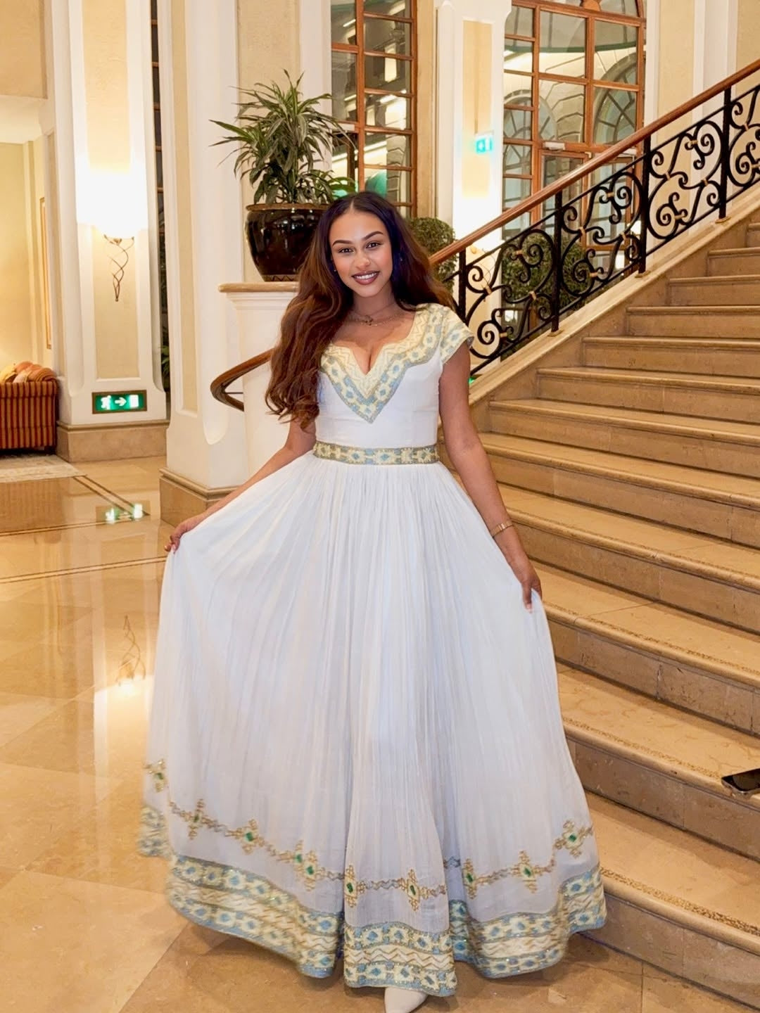 Woman in a white Ethiopian dress with gold embroidery standing in a elegant interior setting.