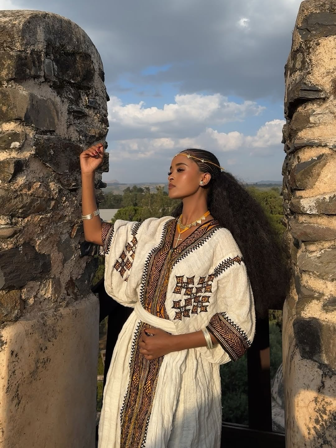 Woman in traditional Gonder Fetel attire standing between stone pillars with a scenic background