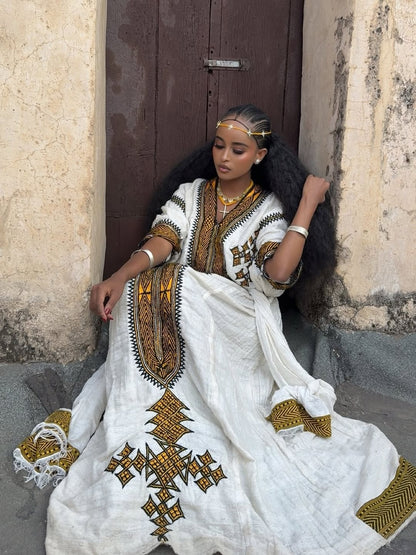 Woman in a white and gold traditional Gonder fetel sitting against a textured wall.