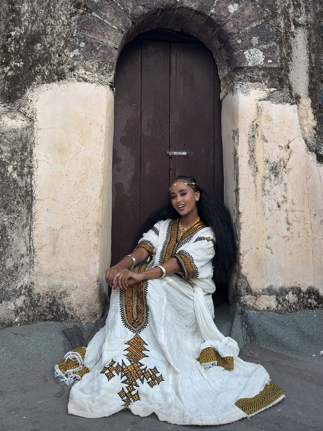 Woman in a white and gold traditional Gonder Fetel sitting in front of a stone archway.