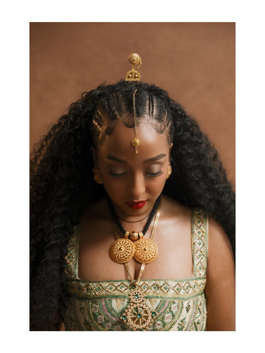 Woman wearing gold jewelry and a green habesha kemis against a brown background