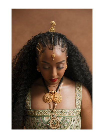 Woman wearing gold jewelry and a green habesha kemis against a brown background