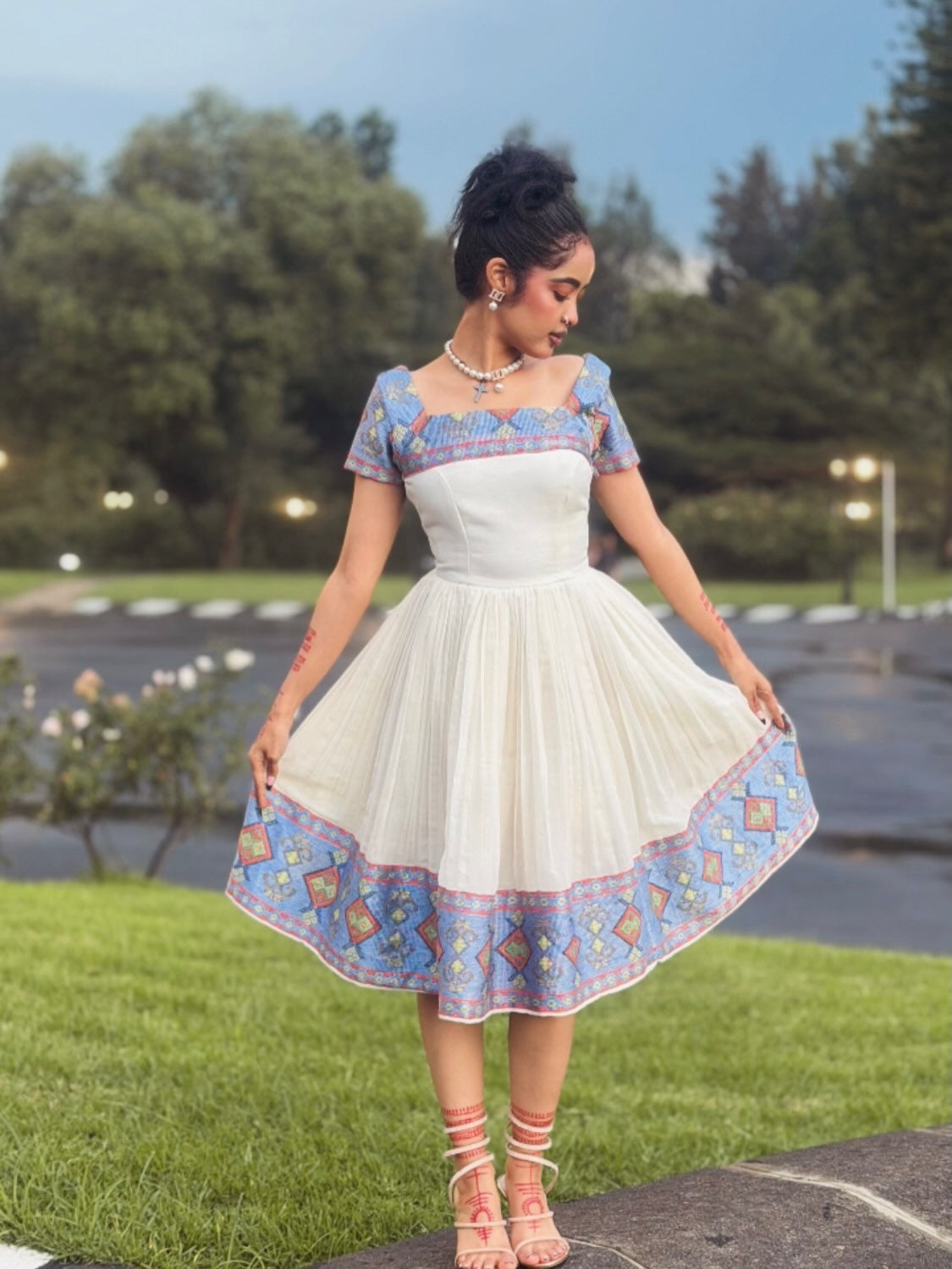 A woman wearing a white A-line Ethiopian Store Dress with blue and orange geometric embroidered borders and a matching embroidered bodice, standing outdoors on a grassy lawn.