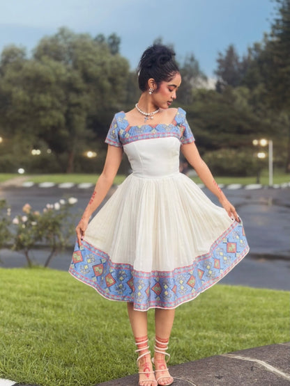 A woman wearing a white A-line Ethiopian Store Dress with blue and orange geometric embroidered borders and a matching embroidered bodice, standing outdoors on a grassy lawn.