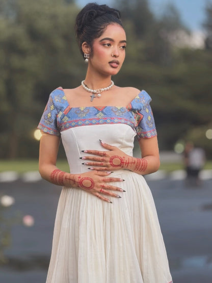 A woman wearing a traditional-style Ethiopian Store Dress with a white skirt and a blue patterned bodice, standing outdoors with her hands decorated with red henna.