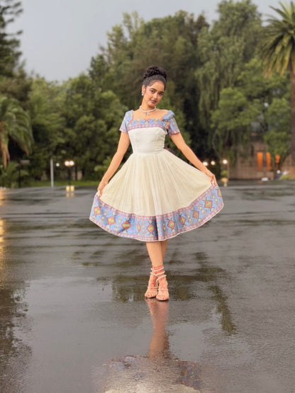 A woman stands on a wet paved path outdoors, twirling in a short cream Ethiopian Store Dress with blue and pink patterned sleeves and hem. Trees and a building are visible in the background.