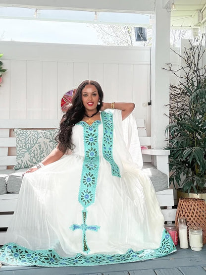 Woman in a white Habesha Dress with turquoise embroidery sitting on a porch.