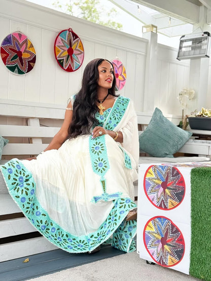 Woman in a white and green saree sitting on a bench with decorative wall art in the background.