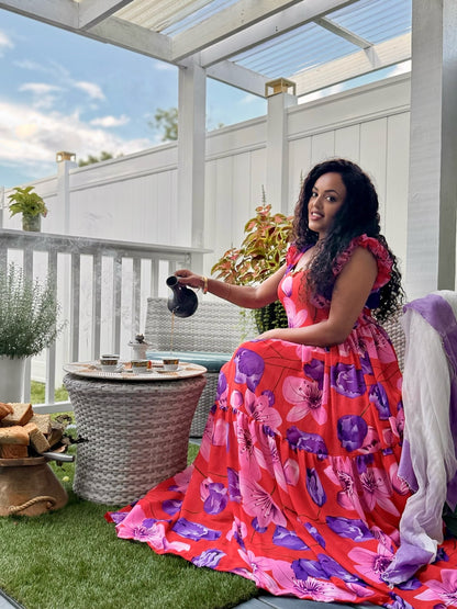 A woman wearing a vibrant pink and purple floral tiered habesha chiffon  sits on a white patio porch, pouring tea from a black teapot into a small woven table teapot.