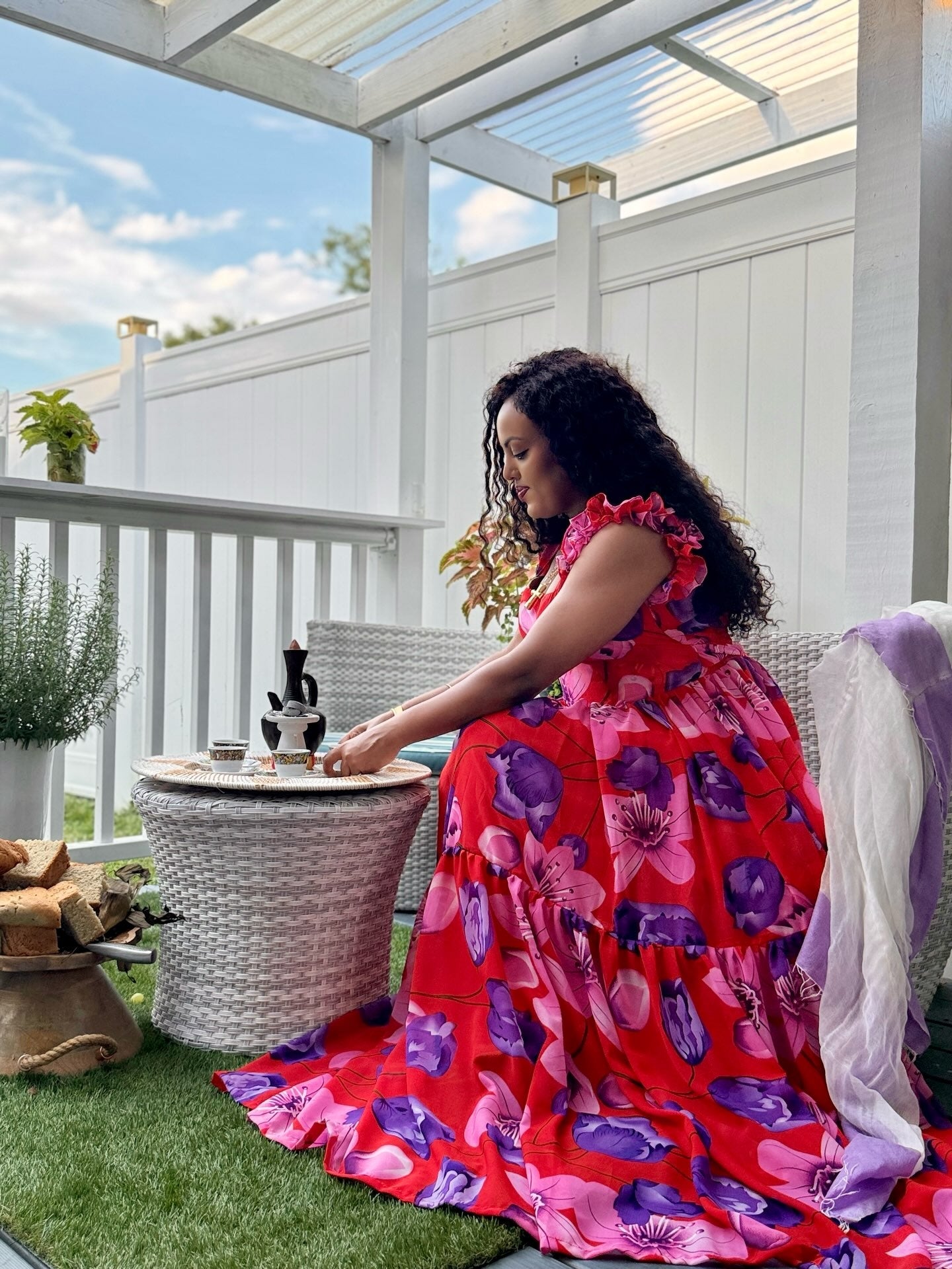 A woman sits on a white wicker patio chair on a backyard porch, wearing a vibrant red tiered habesha chiffon  with a bold purple and pink floral print. She is positioned next to a small wicker side table with a wine bottle and glasses, with a white fence and green grass in the background.
