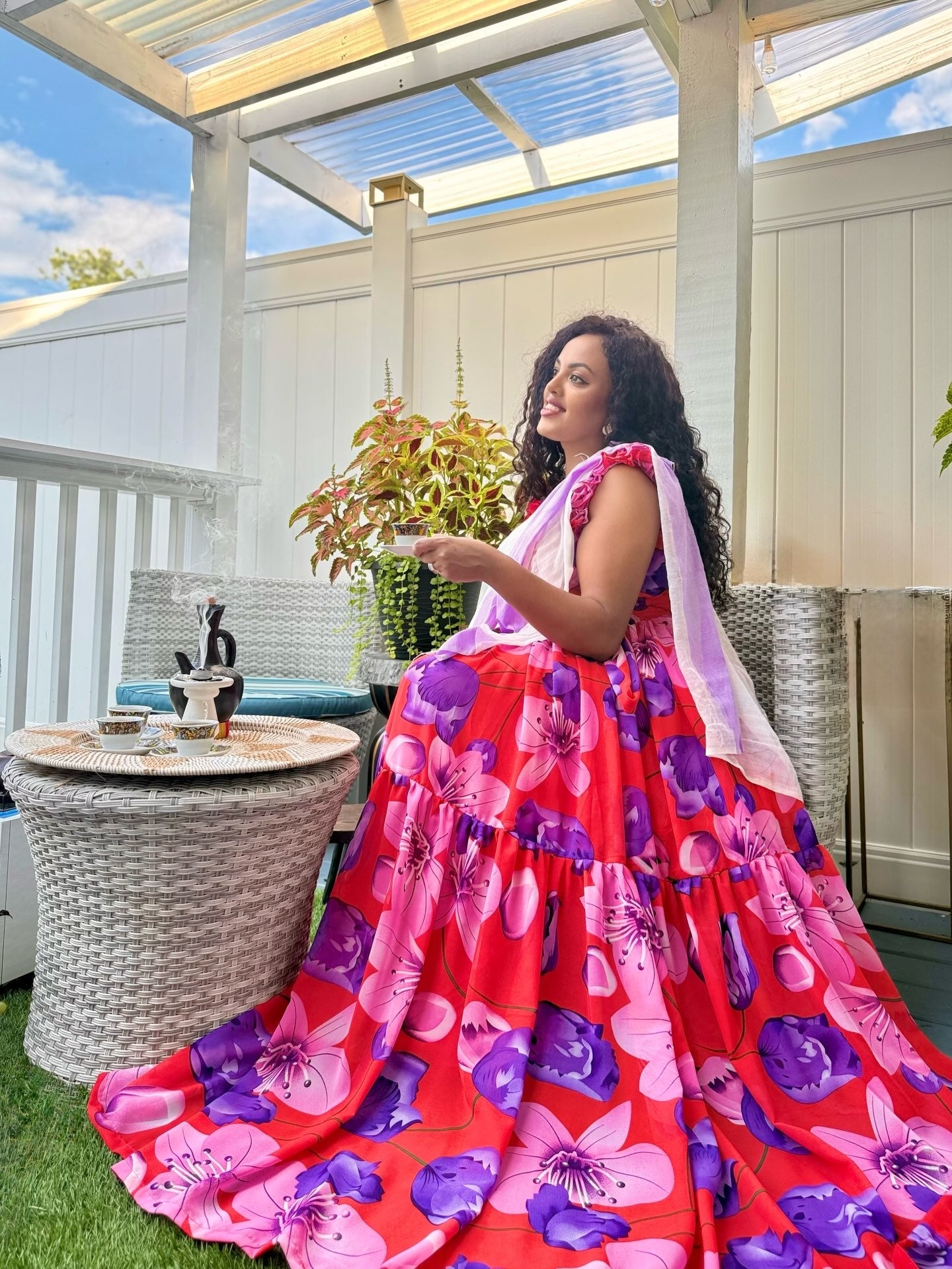 A woman wearing a vibrant red, pink, and purple floral tiered habesha chiffon with a light pink dupatta, posing on a white-walled outdoor patio with wicker furniture.