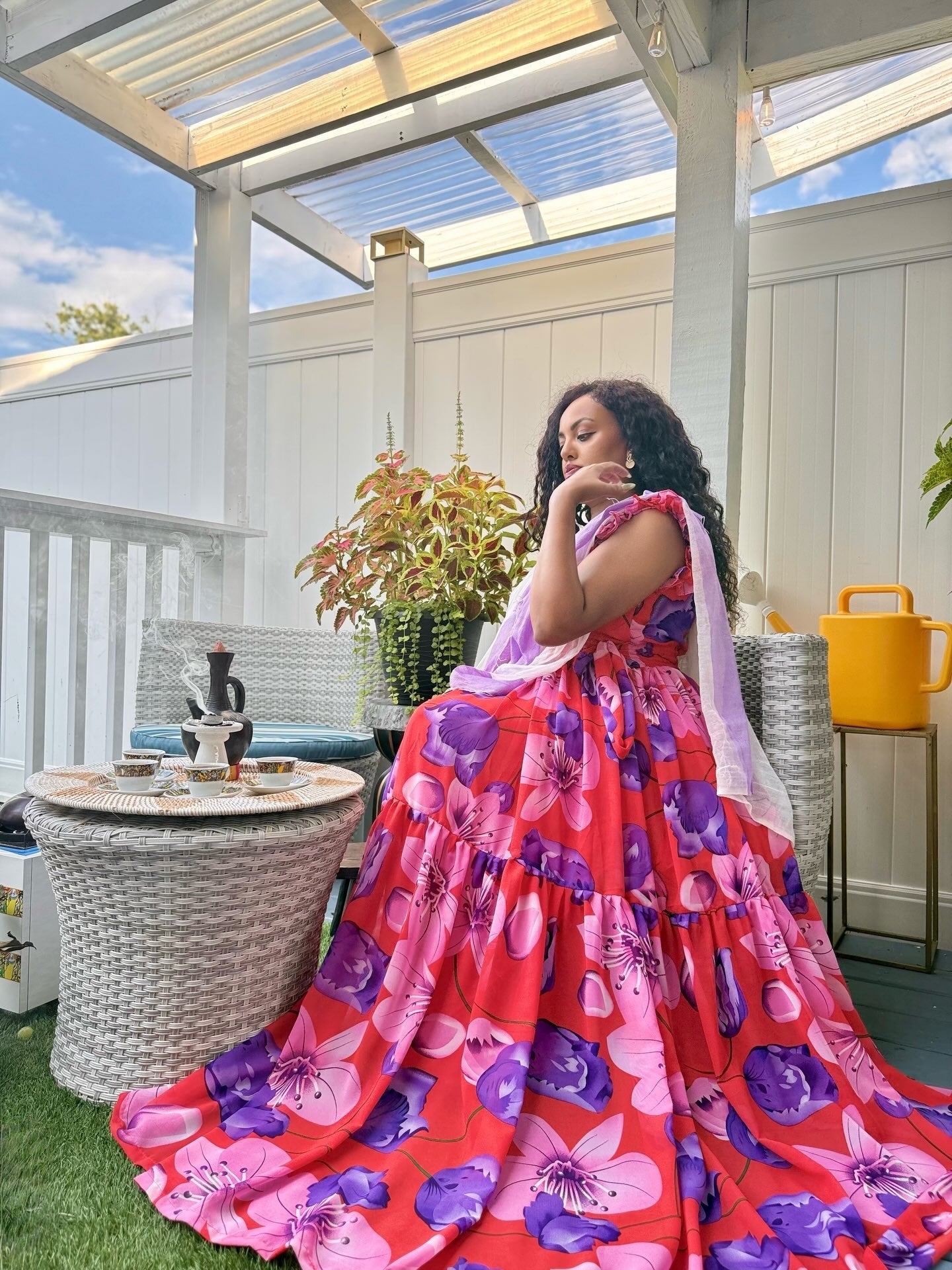 A woman wearing a vibrant tiered ball habesha chiffon with a pink, purple, and red floral print sits on a white wicker patio chair on a covered outdoor porch with a white fence background.