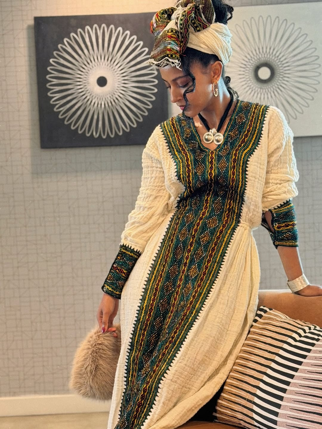 Woman wearing a traditional outfit with intricate patterns in a room with decorative wall art.