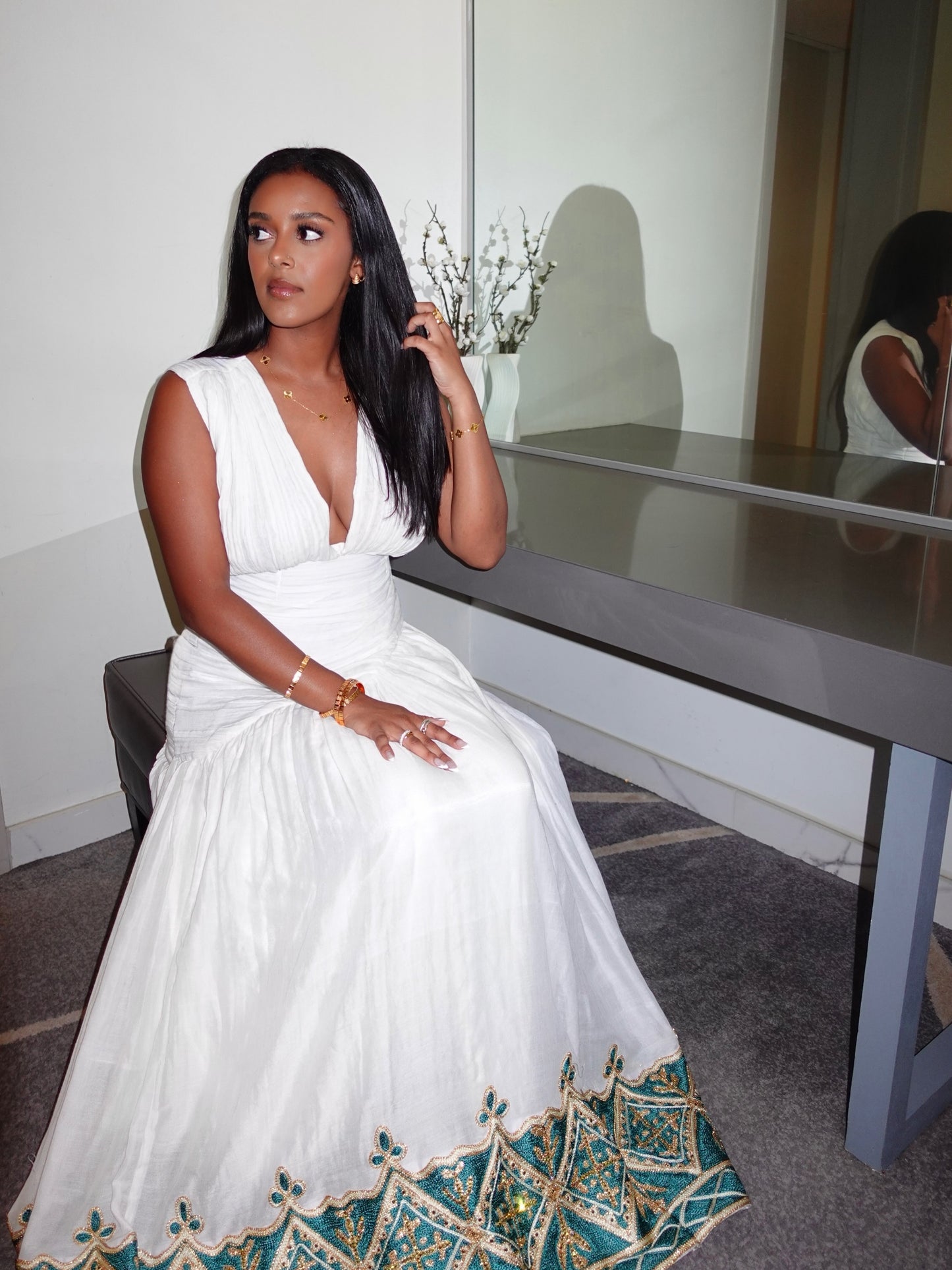 A woman wearing a white sleeveless Habesha Kemis with a deep V-neck and a teal and gold patterned hem, seated in front of a mirror in an indoor setting.