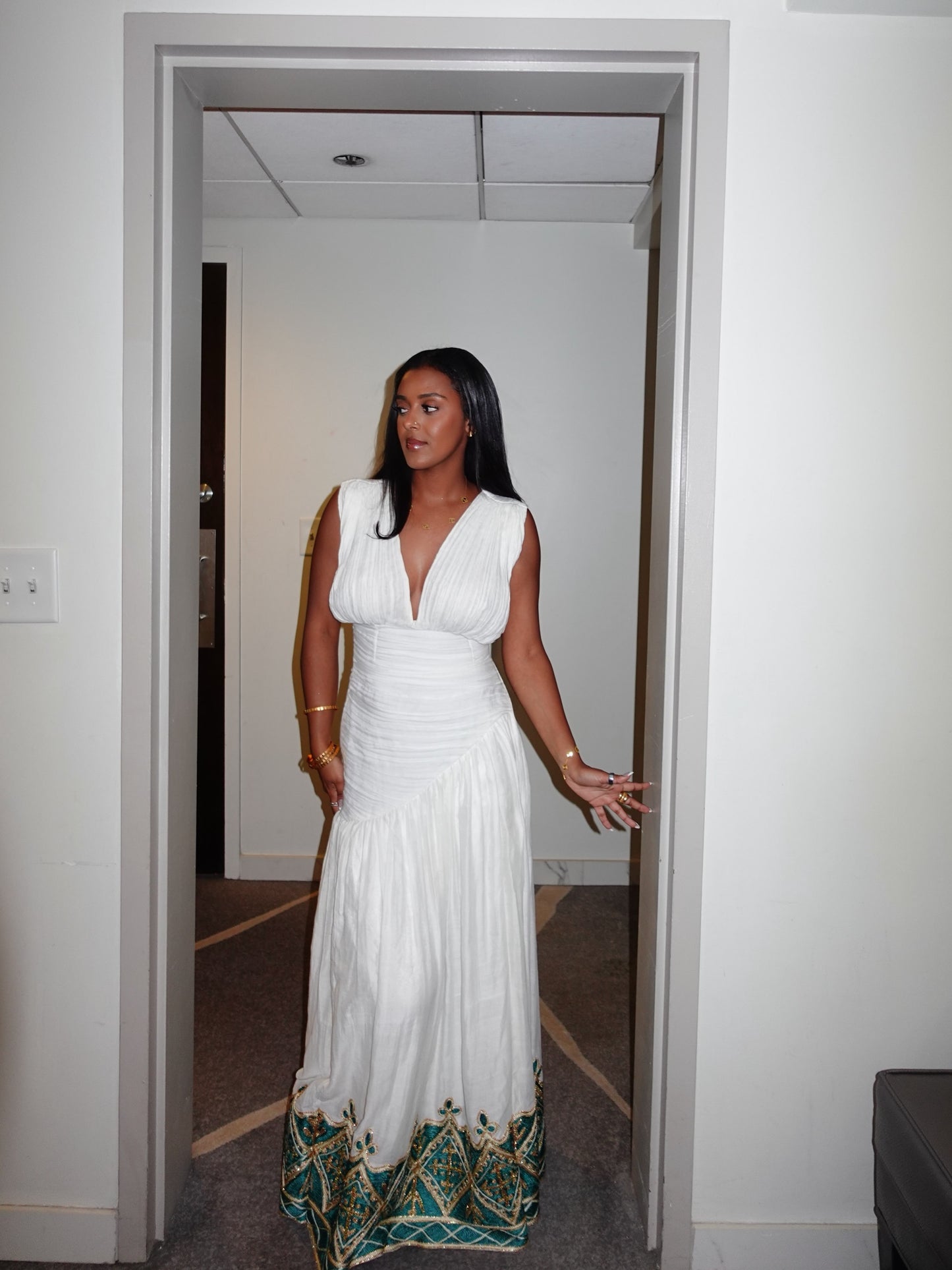 A woman wearing a sleeveless white maxi Habesha Kemis with a deep V-neckline and a decorative teal and gold patterned hem, standing in a doorway.