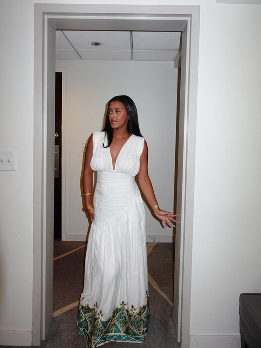 A woman wearing a sleeveless white maxi Habesha Kemis with a deep V-neckline and a decorative teal and gold patterned hem, standing in a doorway.
