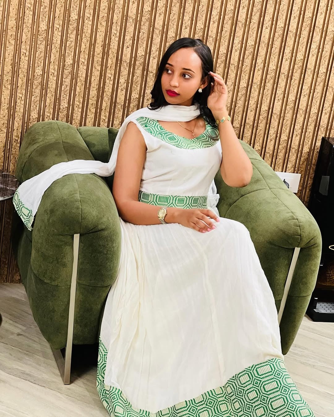 Woman in a white and green traditional outfit sitting on a green chair.