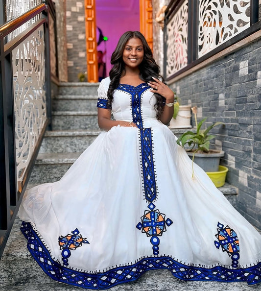 Woman in a white dress with blue embroidery standing outdoors.