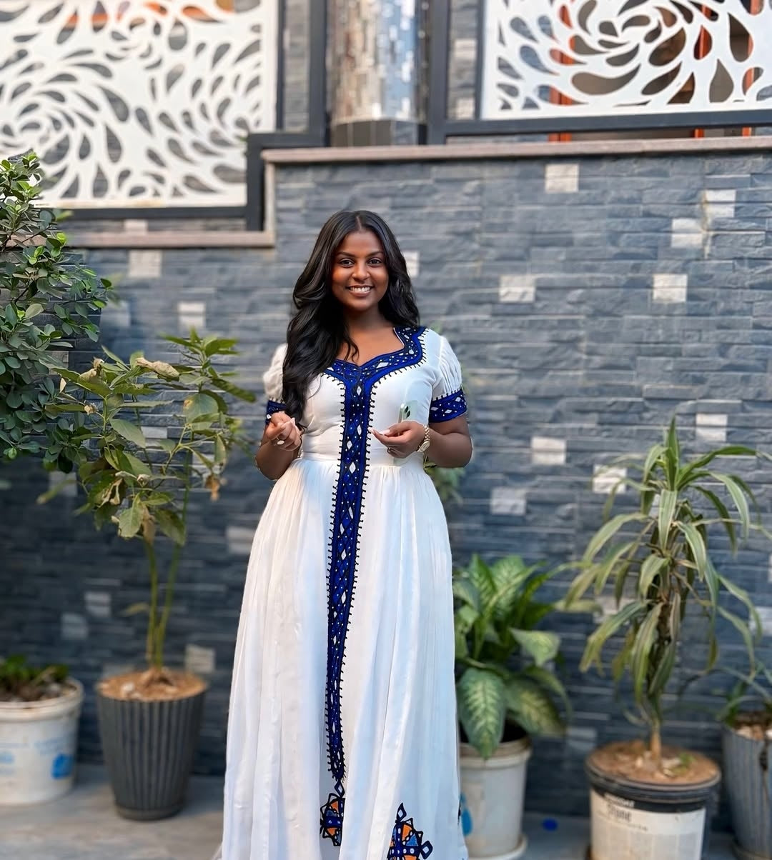 Woman in a white and blue dress standing in front of a decorative wall with plants.