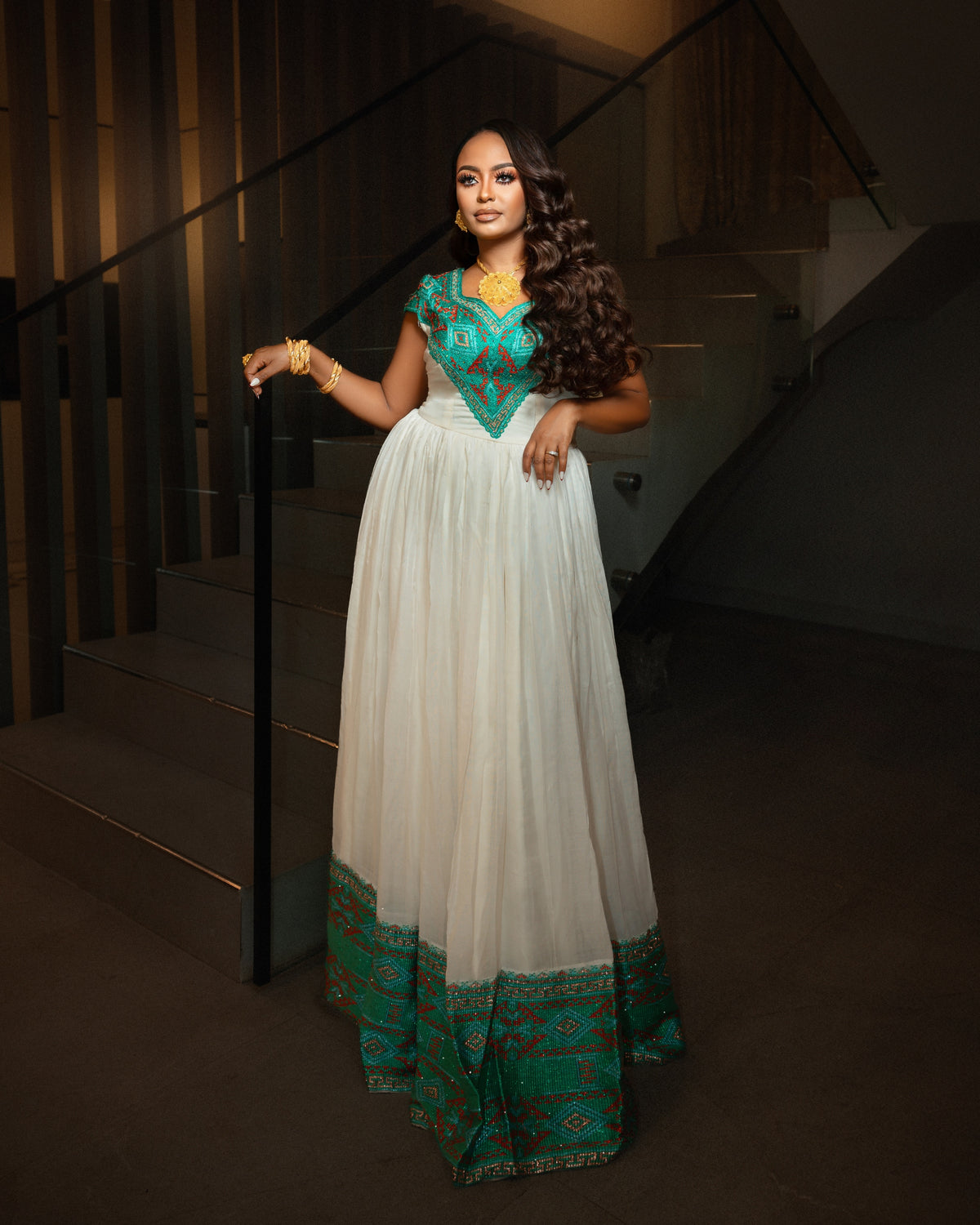A woman wearing a long white  Ethiopian clothing  with a teal and gold embroidered bodice and border, standing in a dimly lit indoor staircase area.