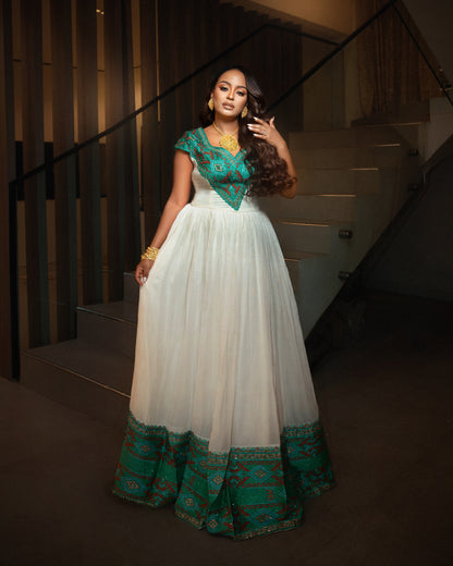 A woman models a traditional  Ethiopian clothing with a white tulle skirt and a teal green embroidered bodice and hem, standing in front of a staircase.