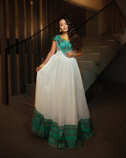 A woman models a traditional Indian  Ethiopian clothing  with a white tulle skirt, a teal green embroidered bodice, and a matching green patterned border at the hem, set against a dimly lit indoor staircase background.