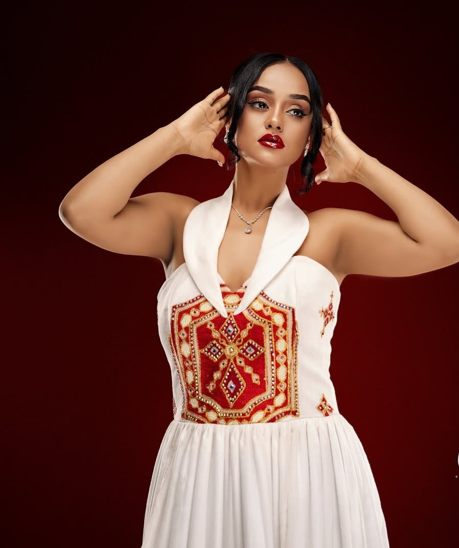 Woman wearing a white dress with red and gold Ethiopian cultural dress embroidery against a dark background